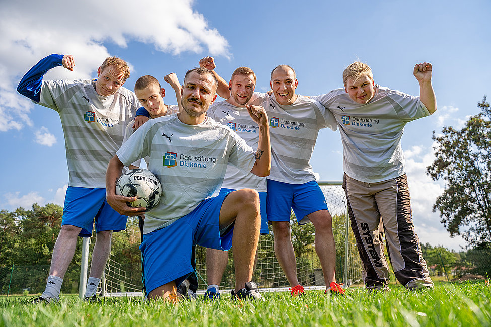 Sechs Männer in Sportbekleidung sind nebeneinander auf einem Fußballplatz, einer der Männer hockt im Vordergrund und hält einen Fußball in der Hand.